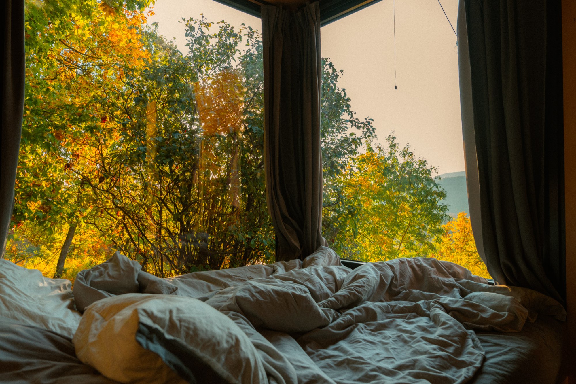 Unmade bed framed by curtains with mist over the hills outside